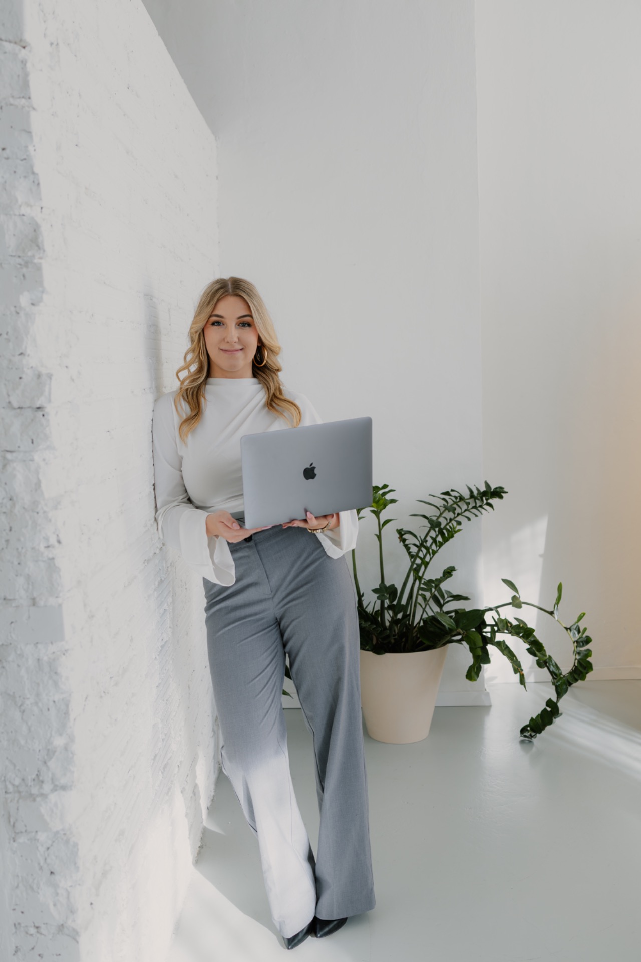 Usměvavá žena v bílé halence drží notebook u bílé zdi, vedle květináč s rostlinou.